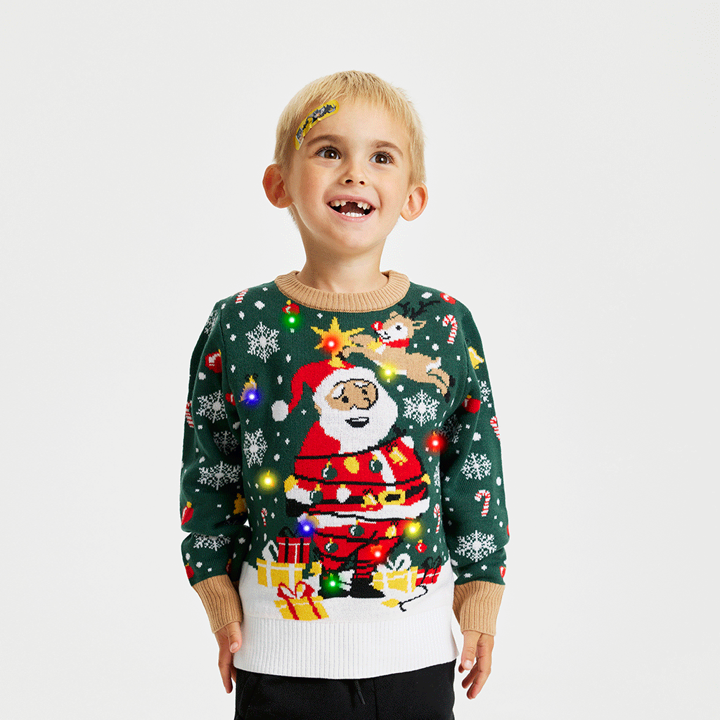 Un enfant souriant portant un pull de Noël vert et blanc avec le Père Noël et des lumières.